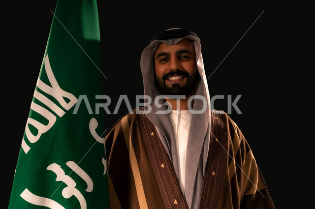 ذكرى تأسيس الدولة السعودية الأولى 1727م، الفخر والاعتزاز بإنجازات المملكة، يوم بدينا 22 فبراير، إظهار الولاء والحب للوطن, بورتريه مقرب لرجل عربي خليجي سعودي يرتدي الغترة والبشت يحمل علم التأسيس ينظر إلى الكاميرا بإيماءات الشموخ، خلفية سوداء