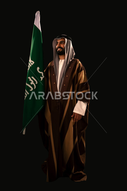 Pride and honor in the achievements of the Kingdom, the day we started February 22, showing loyalty and love for the homeland, the anniversary of the founding of the first Saudi state 1727 AD, a portrait of a Saudi Gulf Arab man wearing a ghutra and a bisht holding the founding flag looking at something with gestures of pride, full body, black background