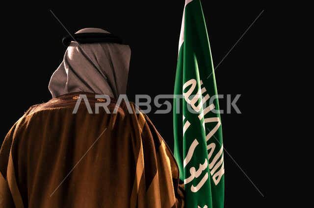 يوم بدينا 22 فبراير، ذكرى تأسيس الدولة السعودية الأولى 1727م، بورتريه مقرب من الخلف لرجل عربي خليجي سعودي يرتدي الغترة والبشت يحمل علم التأسيس، إظهار الولاء والحب للوطن, الفخر والاعتزاز بإنجازات المملكة، خلفية سوداء
