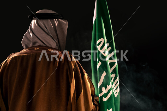 ذكرى تأسيس الدولة السعودية الأولى 1727م، بورتريه مقرب من الخلف لرجل عربي خليجي سعودي يرتدي الغترة والبشت يحمل علم التأسيس، إظهار الولاء والحب للوطن, الفخر والاعتزاز بإنجازات المملكة، يوم بدينا 22 فبراير، خلفية سوداء