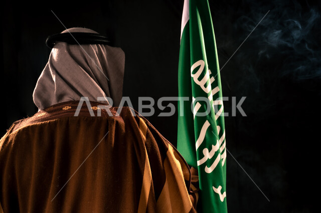 يوم بدينا 22 فبراير، ذكرى تأسيس الدولة السعودية الأولى 1727م، بورتريه مقرب من الخلف لرجل عربي خليجي سعودي يرتدي الغترة والبشت يحمل علم التأسيس، إظهار الولاء والحب للوطن, الفخر والاعتزاز بإنجازات المملكة، خلفية سوداء