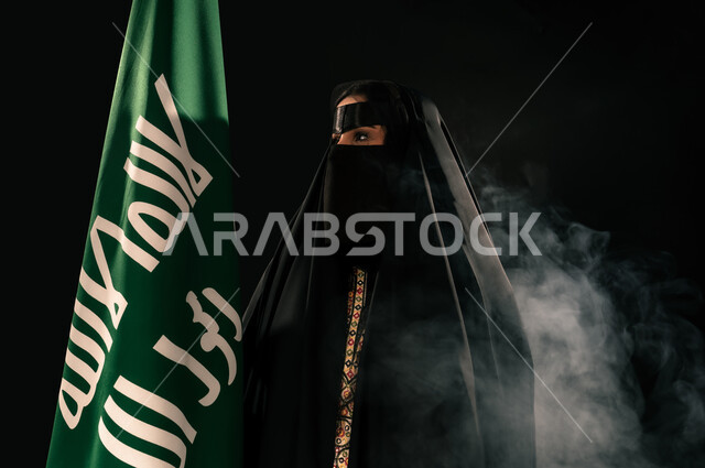 ذكرى تأسيس الدولة السعودية الأولى 1727م، بورتريه مقرب لامرأة عربية خليجية سعودية ترتدي البرقع والعباءة تحمل علم التأسيس تنظر إلى شيء ما بإيماءات الاعتزاز والشموخ، إظهار الولاء والحب للوطن, الفخر بإنجازات المملكة، يوم بدينا 22 فبراير، خلفية سوداء