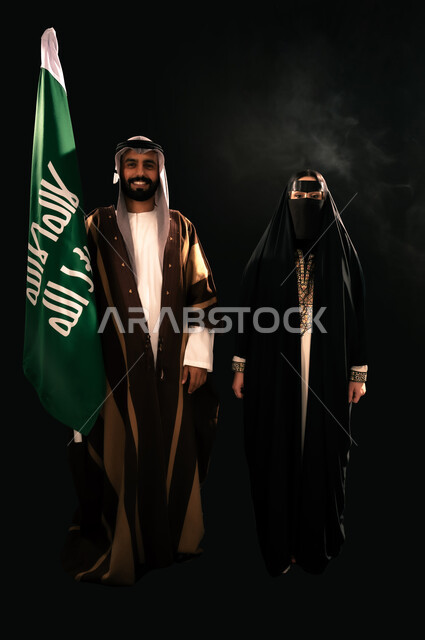 إظهار الولاء والحب للوطن, ذكرى تأسيس الدولة السعودية الأولى 1727م، يوم بدينا 22 فبراير، بورتريه مقرب لرجل وامرأة عربيين خليجيين سعوديين يرتديان الزي الشعبي التقليدي يقفان بجانب علم التأسيس ينظران إلى الكاميرا بإيماءات الاعتزاز، الجسم بالطول الكامل، خلفية سوداء