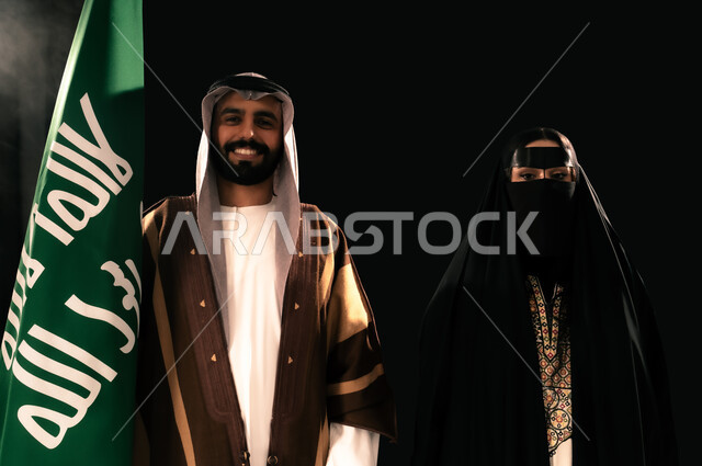ذكرى تأسيس الدولة السعودية الأولى 1727م، يوم بدينا 22 فبراير، الثقة والفخر بإنجازات المملكة، بورتريه مقرب لرجل وامرأة عربيين خليجيين سعوديين يرتديان الزي الشعبي التقليدي يقفان بجانب علم التأسيس ينظران إلى الكاميرا بإيماءات الاعتزاز، إظهار الولاء والحب للوطن, خلفية سوداء