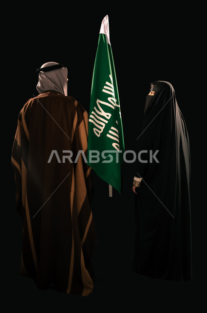 ذكرى تأسيس الدولة السعودية الأولى 1727م، إظهار الولاء والحب للوطن, الثقة والفخر بإنجازات المملكة، بورتريه من الخلف لرجل وامرأة عربيين خليجيين سعوديين يرتديان الزي الشعبي التقليدي يقفان بجانب علم التأسيس، يوم بدينا 22 فبراير، الجسم بالطول الكامل، خلفية سوداء