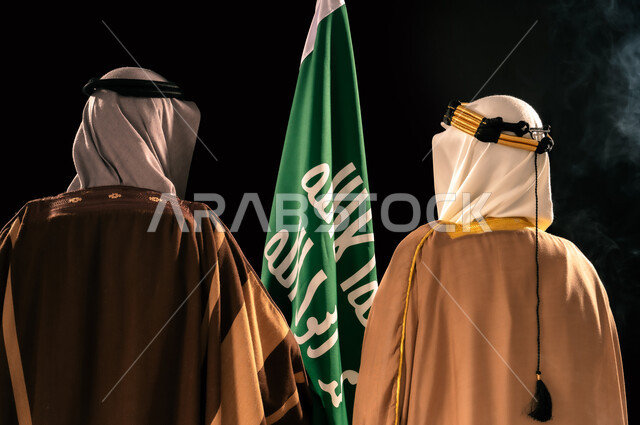 يوم بدينا 22 فبراير، الثقة والفخر بإنجازات المملكة، ذكرى تأسيس الدولة السعودية الأولى 1727م، إظهار الولاء والحب للوطن, بورتريه مقرب من الخلف لرجلين عربيين خليجيين سعوديين يرتدان الزي الشعبي التقليدي يقفان بجانب علم التأسيس، خلفية سوداء