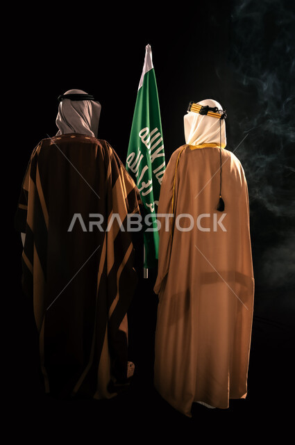 ذكرى تأسيس الدولة السعودية الأولى 1727م، إظهار الولاء والحب للوطن, الثقة والفخر بإنجازات المملكة، بورتريه من الخلف لرجلين عربيين خليجيين سعوديين يرتدان الزي الشعبي التقليدي يقفان بجانب علم التأسيس، يوم بدينا 22 فبراير، الجسم بالطول الكامل، خلفية سوداء