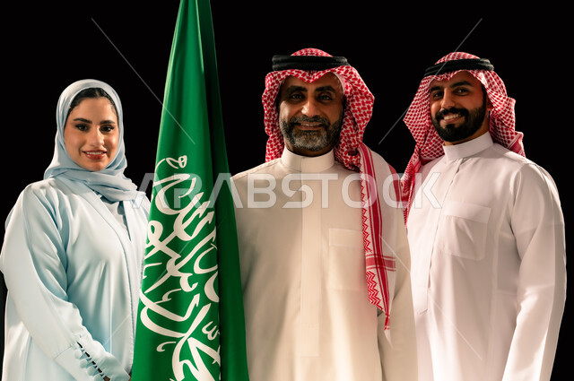 النظر الى الكاميرا بإيماءات السرور و الفخر, يوم العلم 11 مارس, ترسيخ جذور الانتماء الوطني, إحياء ذكرى اليوم الوطني السعودي 23 سبتمبر, اظهار الحب و التفاني للبلاد, بورتريه مقرب لعائلة عربية خليجية سعودية مبتسمة تقف بجانب علم المملكة, خلفية سوداء