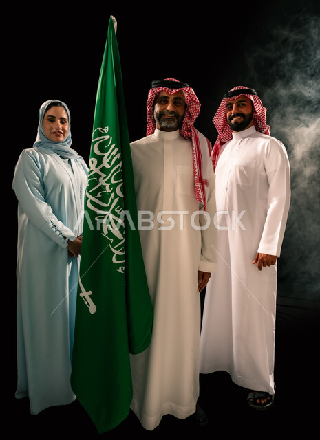 اظهار الحب و التفاني للبلاد, النظر الى الكاميرا بإيماءات الفخر, يوم العلم 11 مارس, إحياء ذكرى اليوم الوطني السعودي 23 سبتمبر, ترسيخ جذور الانتماء الوطني, بورتريه لعائلة عربية خليجية سعودية مبتسمة تقف بجانب علم المملكة, صورة الجسم بالطول الكامل, خلفية سوداء