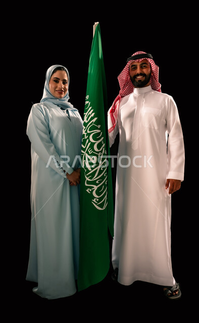 النظر الى الكاميرا بإيماءات الفخر و السرور, يوم العلم 11 مارس, إحياء ذكرى اليوم الوطني السعودي 23 سبتمبر, ترسيخ جذور الانتماء الوطني, بورتريه لزوجان عربيان خليجيان سعوديان مبتسمان يقفان بجانب علم المملكة, صورة الجسم بالطول الكامل, خلفية سوداء