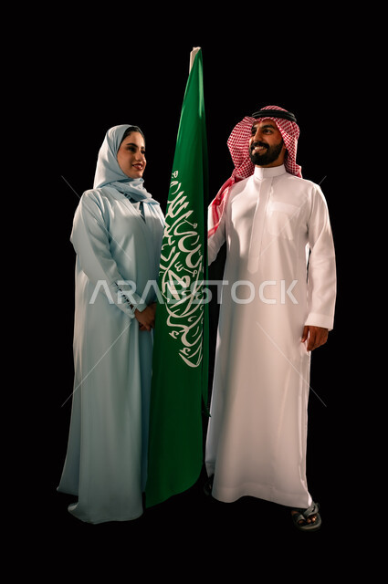 يوم العلم 11 مارس, إحياء ذكرى اليوم الوطني السعودي 23 سبتمبر, ترسيخ جذور الانتماء الوطني, النظر الى العلم بإيماءات الفخر و السرور, بورتريه لزوجان عربيان خليجيان سعوديان مبتسمان يقفان بجانب علم المملكة, صورة الجسم بالطول الكامل, خلفية سوداء