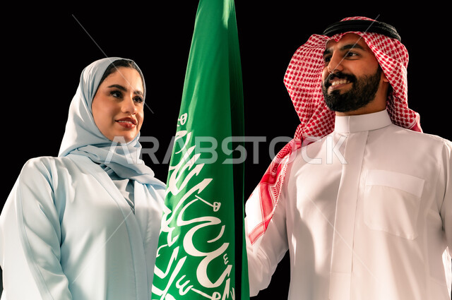 ترسيخ جذور الانتماء الوطني, النظر الى العلم بإيماءات الفخر و السرور, إحياء ذكرى اليوم الوطني السعودي 23 سبتمبر, يوم العلم 11 مارس, بورتريه مقرب لشاب و امرأة عربيان خليجيان سعوديان مبتسمان يقفان بجانب علم المملكة, خلفية سوداء