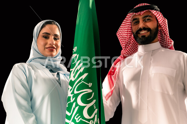 إحياء ذكرى اليوم الوطني السعودي 23 سبتمبر, يوم العلم 11 مارس, ترسيخ جذور الانتماء الوطني, النظر الى الكاميرا بإيماءات الفخر و السرور, بورتريه مقرب لشاب و امرأة عربيان خليجيان سعوديان مبتسمان يقفان بجانب علم المملكة, خلفية سوداء