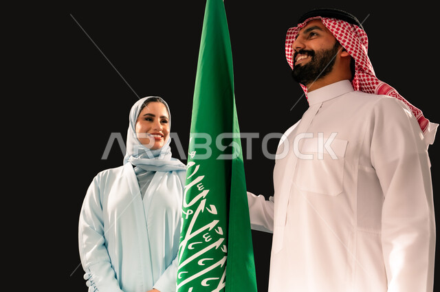 ترسيخ جذور الانتماء الوطني, النظر الى العلم بإيماءات الفخر و السرور, إحياء ذكرى اليوم الوطني السعودي 23 سبتمبر, يوم العلم 11 مارس, بورتريه مقرب لشاب و امرأة عربيان خليجيان سعوديان مبتسمان يقفان بجانب علم المملكة, خلفية سوداء