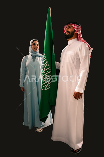 النظر الى العلم بإيماءات الفخر و السرور بالانتماء للوطن, إحياء ذكرى اليوم الوطني السعودي 23 سبتمبر, يوم العلم 11 مارس, بورتريه لشاب و امرأة عربيان خليجيان سعوديان مبتسمان يقفان بجانب علم المملكة, صورة الجسم بالطول الكامل, خلفية سوداء