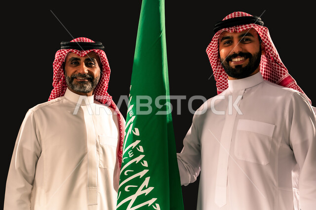 النظر الى الكاميرا بإيماءات الفخر و السرور بالانتماء للوطن, إحياء ذكرى اليوم الوطني السعودي 23 سبتمبر, يوم العلم 11 مارس, بورتريه مقرب لرجلان عربيان خليجيان سعوديان مبتسمان يرتديان الشماغ و الثوب التقليدي يقفان بجانب علم المملكة, خلفية سوداء