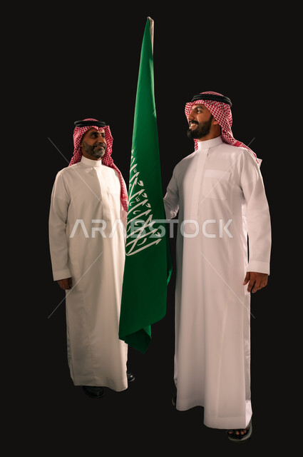 ذكرى اليوم الوطني السعودي 23 سبتمبر، مفهوم حب الوطن والفخر بالانتماء الى البلاد، بورتريه لرجلين عربيين خليجيين سعوديين يرتديان الثوب التقليدي والشماغ يحملان علم المملكة ينظران الى بعضهما بتعابير السرور، صورة الجسم بالطول الكامل، يوم العلم 11 مارس، خلفية سوداء