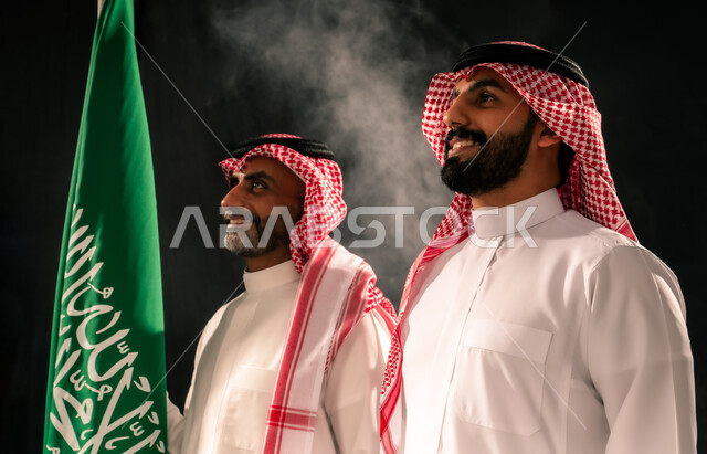 ذكرى اليوم الوطني السعودي 23 سبتمبر، مفهوم حب الوطن والفخر بالانتماء الى البلاد، بورتريه مقرب لرجلين عربيين خليجيين سعوديين يرتديان الثوب التقليدي والشماغ ينظران الى شيء ما بتعابير السرور، رجل سعودي يحمل علم المملكة، يوم العلم 11 مارس، خلفية سوداء
