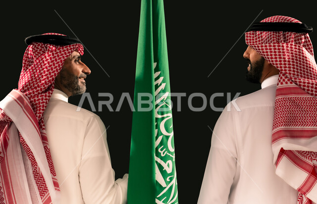 ذكرى اليوم الوطني السعودي 23 سبتمبر، بورتريه مقرب من الخلف لرجلين عربيين خليجيين سعوديين يرتديان الثوب التقليدي والشماغ يمسكان علم المملكة ينظران لبعضهما، مفهوم حب الوطن والفخر بالانتماء الى البلاد، يوم العلم 11 مارس، خلفية سوداء