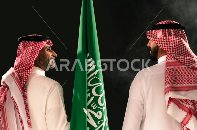 ذكرى اليوم الوطني السعودي 23 سبتمبر، بورتريه مقرب من الخلف لرجلين عربيين خليجيين سعوديين يرتديان الثوب التقليدي والشماغ يمسكان علم المملكة ينظران لبعضهما، مفهوم حب الوطن والفخر بالانتماء الى البلاد، يوم العلم 11 مارس، خلفية سوداء