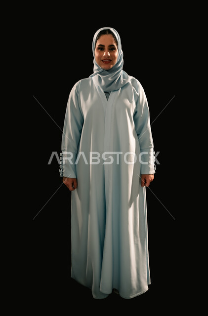 Concern for elegance and appearance, concept of femininity and softness, confidence and self-esteem, portrait of a smiling veiled Saudi Arabian Gulf woman wearing a blue abaya standing straight looking at the camera with gestures of pleasure, full-length body image, black background