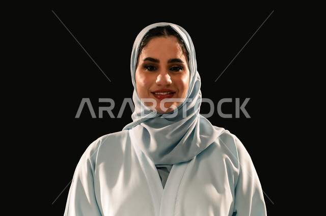 The concept of femininity and softness, interest in elegance and appearance, confidence and self-esteem, close-up portrait of a smiling veiled Saudi Arabian Gulf woman wearing a blue abaya looking at the camera with gestures of pleasure, black background