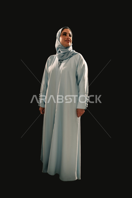 Confidence and self-esteem, interest in elegance and appearance, concept of femininity and softness, portrait of a smiling veiled Saudi Arabian Gulf woman wearing a blue abaya looking at something with gestures of pleasure, full-length body image, black background