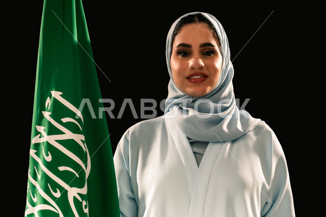 النظر الى الكاميرا بتعابير السرور, يوم العلم 11 مارس، إحياء ذكرى اليوم الوطني السعودي 23 سبتمبر, الاعتزاز و الفخر بالانتماء للوطن, رفع راية التوحيد والإسلام، بورتريه مقرب لامرأة عربية خليجية سعودية مبتسمة محجبة ترتدي العباءه تحمل بيدها علم المملكة، خلفية سوداء