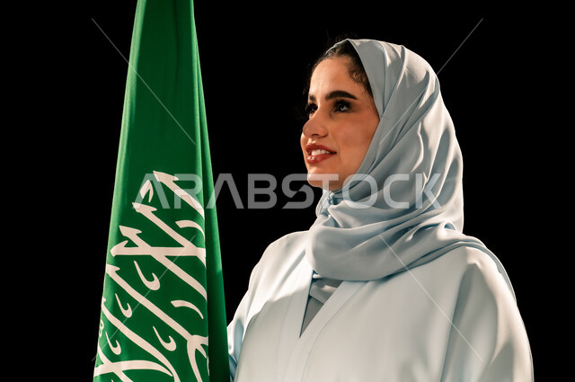 إحياء ذكرى اليوم الوطني السعودي 23 سبتمبر, النظر الى شيء ما بتعابير السرور, الاعتزاز و الفخر بالانتماء للوطن, رفع راية التوحيد والإسلام، يوم العلم 11 مارس، بورتريه مقرب لامرأة عربية خليجية سعودية مبتسمة محجبة ترتدي العباءه تحمل بيدها علم المملكة، خلفية سوداء