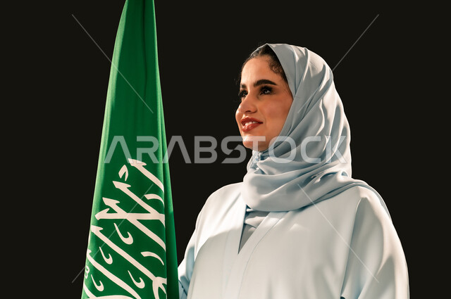 النظر الى شيء ما بتعابير السرور, الاعتزاز و الفخر بالانتماء للوطن, رفع راية التوحيد والإسلام، يوم العلم 11 مارس، إحياء ذكرى اليوم الوطني السعودي 23 سبتمبر, بورتريه مقرب لامرأة عربية خليجية سعودية مبتسمة محجبة ترتدي العباءه تحمل بيدها علم المملكة، خلفية سوداء