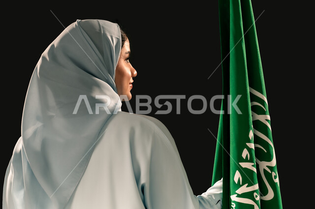 رفع راية التوحيد والإسلام، يوم العلم 11 مارس، إحياء ذكرى اليوم الوطني السعودي 23 سبتمبر, بورتريه مقرب من الخلف لامرأة عربية خليجية سعودية مبتسمة محجبة ترتدي العباءه تحمل بيدها علم المملكة وتنظر الى شيء ما، الفخر و الاعتزاز بالانتماء للوطن, خلفية سوداء