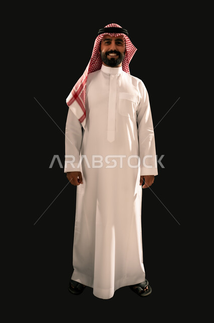 The concept of masculinity and elegance, interest in external appearance, portrait of a smiling Saudi Arabian Gulf young man wearing traditional thobe and shemagh looking at the camera with gestures of joy and happiness, full-length body image, black background
