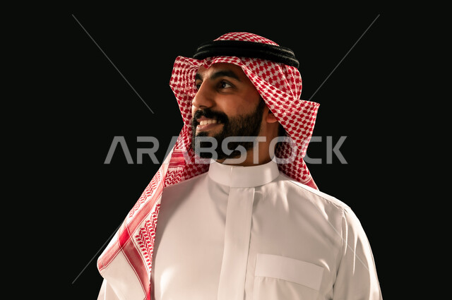 The concept of masculinity and elegance, interest in outward appearance, close-up portrait of a smiling Saudi Arabian Gulf young man wearing traditional thobe and shemagh looking at something with gestures of joy and happiness, black background