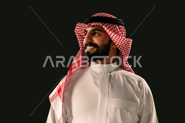 The concept of masculinity and elegance, interest in outward appearance, close-up portrait of a smiling Saudi Arabian Gulf young man wearing traditional thobe and shemagh looking at something with gestures of joy and happiness, black background