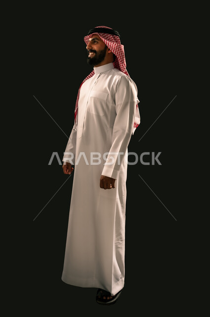 The concept of masculinity and elegance, interest in appearance, side portrait of a smiling Saudi Arabian Gulf young man wearing traditional thobe and shemagh looking at something with gestures of joy and happiness, full body image, black background