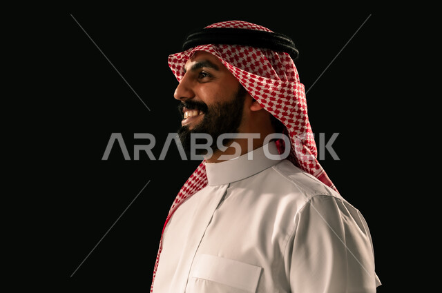 الاهتمام بالمظهر الخارجي، مفهوم الرجولة والأناقة، بورتريه مقرب من الجانب لشاب عربي خليجي سعودي مبتسم يرتدي الثوب التقليدي والشماغ ينظر الى شيء ما بإيماءات الفرح والسرور، خلفية سوداء