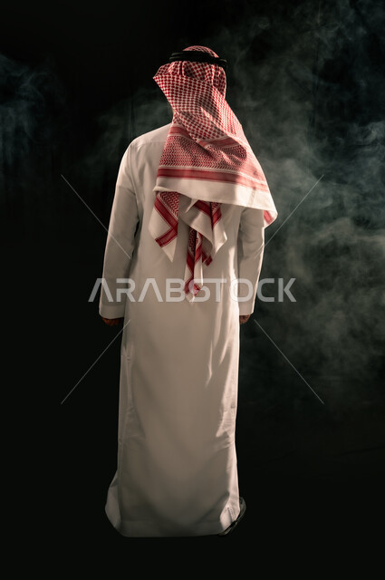 Concept of masculinity and elegance, interest in appearance, portrait from behind of a Saudi Arabian Gulf man wearing traditional thobe and shemagh standing straight, full body image, black background