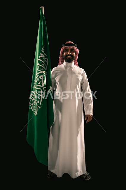 النظر الى الكاميرا بإيماءات الفخر و السرور بالانتماء للوطن, إحياء ذكرى اليوم الوطني السعودي 23 سبتمبر, يوم العلم 11 مارس, بورتريه لشاب عربي خليجي سعودي مبتسم يرتدي الشماغ و الثوب التقليدي يحمل بيده علم المملكة, صورة الجسم بالطول الكامل, خلفية سوداء