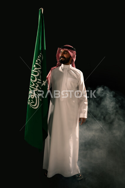 يوم العلم 11 مارس, النظر الى العلم بإيماءات الفخر و السرور بالانتماء للوطن, إحياء ذكرى اليوم الوطني السعودي 23 سبتمبر, بورتريه لشاب عربي خليجي سعودي مبتسم يرتدي الشماغ و الثوب التقليدي يحمل بيده علم المملكة, صورة الجسم بالطول الكامل, خلفية سوداء
