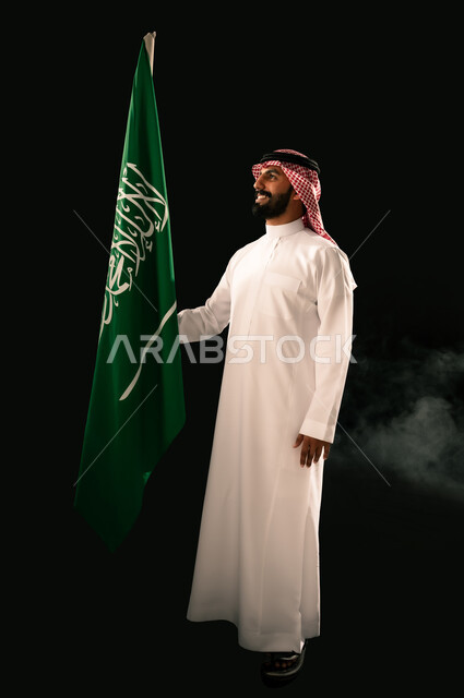 النظر الى العلم بإيماءات الفخر و السرور بالانتماء للوطن, إحياء ذكرى اليوم الوطني السعودي 23 سبتمبر, يوم العلم 11 مارس, بورتريه لشاب عربي خليجي سعودي مبتسم يرتدي الشماغ و الثوب التقليدي يحمل بيده علم المملكة, صورة الجسم بالطول الكامل, خلفية سوداء
