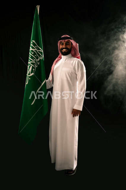 النظر الى الكاميرا بإيماءات الفخر بالانتماء للوطن, الاهتمام بالمظهر الخارجي, يوم العلم 11 مارس, بورتريه لرجل عربي خليجي سعودي مبتسم يرتدي الشماغ و الثوب التقليدي يحمل بيده علم المملكة, إحياء ذكرى اليوم الوطني السعودي 23 سبتمبر, الجسم بالطول الكامل, خلفية سوداء