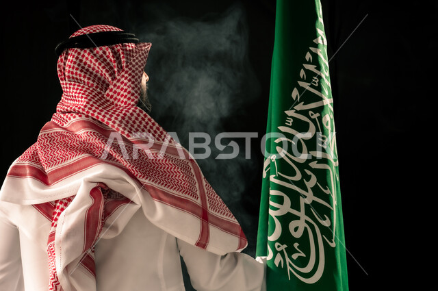 الاهتمام بالمظهر الخارجي, إحياء ذكرى اليوم الوطني السعودي 23 سبتمبر, يوم العلم 11 مارس, مفهوم حب الوطن و الفخر بالانتماء للبلاد, بورتريه مقرب من الخلف لرجل عربي خليجي سعودي يرتدي الشماغ و الثوب التقليدي ينظر إلى علم المملكة, خلفية سوداء