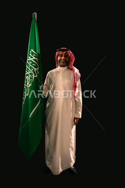 يوم العلم 11 مارس, النظر الى الكاميرا بإيماءات الفخر بالانتماء للوطن, بورتريه لرجل عربي خليجي سعودي مبتسم يرتدي الشماغ و الثوب التقليدي يحمل بيده علم المملكة, إحياء ذكرى اليوم الوطني السعودي 23 سبتمبر, الجسم بالطول الكامل, خلفية سوداء