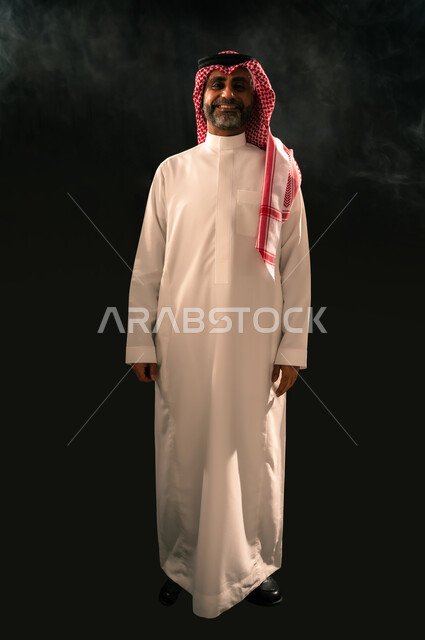The concept of masculinity and self-confidence, interest in elegance and appearance, portrait of a smiling Saudi Arabian Gulf man wearing traditional thobe and shemagh standing straight looking at the camera with gestures of joy and happiness, full body image, black background