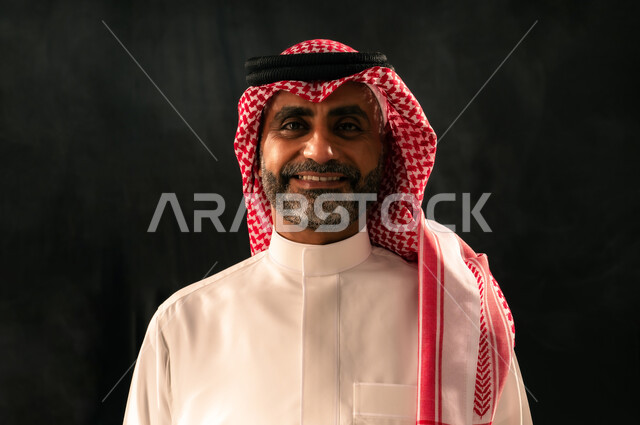 Taking portraits for official documents, concern for appearance, concept of masculinity and elegance, close-up portrait of a smiling Saudi Arabian Gulf man wearing traditional thobe and shemagh standing straight looking at the camera with gestures of joy and happiness, black background
