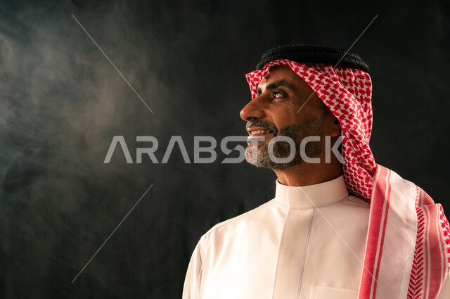 The concept of masculinity and elegance, interest in appearance, close-up portrait from the side of a smiling Saudi Arabian Gulf man wearing traditional thobe and shemagh standing straight looking at something with gestures of joy and happiness, black background