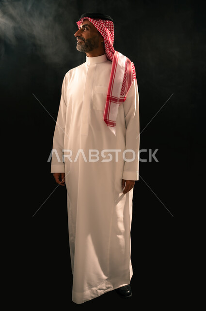 الأناقة و الاهتمام بالمظهر الخارجي، مفهوم الرجولة والثقة بالنفس، بورتريه لرجل عربي خليجي سعودي مبتسم يرتدي الثوب التقليدي والشماغ يقف باستقامة ينظر الى شيء ما بإيماءات الفرح والسرور، صورة الجسم بالطول الكامل، خلفية سوداء