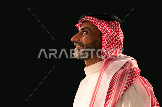 Concern for elegance and appearance, concept of masculinity and self-confidence, close-up side portrait of a smiling Saudi Arabian Gulf man wearing traditional thobe and shemagh looking at something with gestures of joy and happiness, black background