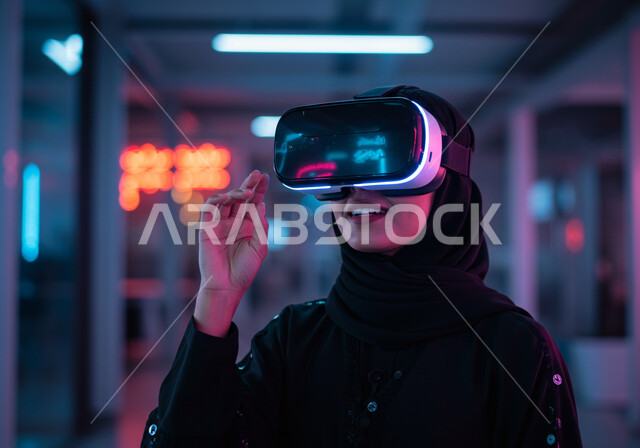 التفاعل والاندماج مع العناصر الهولوجرامية، محاكاه العالم الخيالي، استخدام الأجهزة التقنية الحديثة والمتطورة، صورة مقربة لامرأة عربية خليجية سعودية محجبة ترتدي عباءة سوداء وتضع نظارة الواقع الافتراضي، مفهوم التطور التقني والتكنولوجي، ايماءات محاولة الامساك بشيء ما