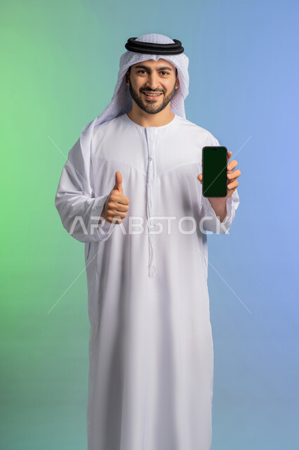 رفع اصبع الابهام للأعلى بايماءات الجودة والامتياز، مفهوم القبول والإعجاب والرضا, عرض تطبيقات الجوال عبر شاشة خضراء فارغة كروما،  بورتريه لرجل عربي خليجي اماراتي يرتدي الكندورة والغترة البيضاء يمسك بيده الهاتف المحمول، استخدام جهاز تقني حديث ومتطور، خلفية ملونة